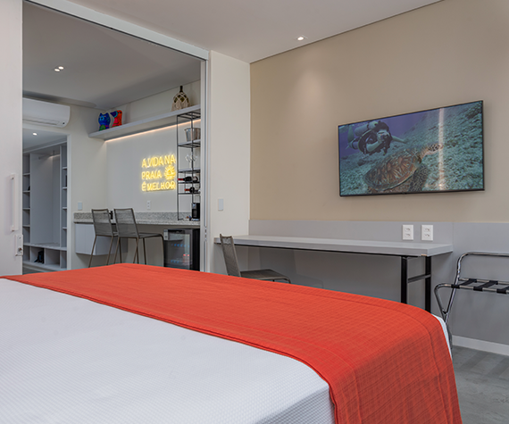   Interior da Garden Suite, com uma cama de casal e uma TV. O ambiente é claro, com um balcão, com algumas cadeiras e um letreiro de neon no fundo.
