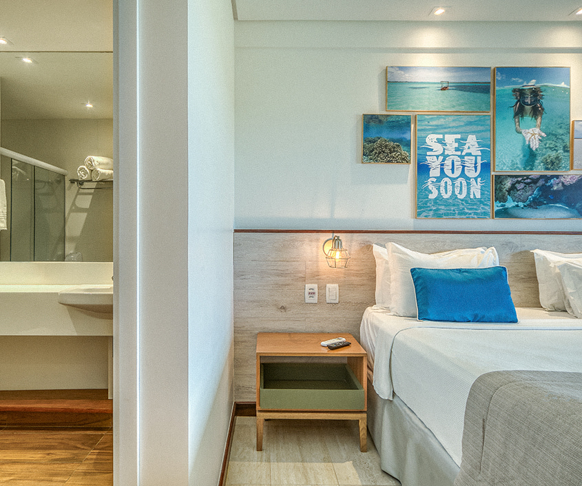  Quarto Standard do Japaratinga Lounge Resort com decoração praiana, exibindo uma cama com travesseiros azuis e quadros temáticos de mar à direita, e a entrada para um banheiro iluminado com bancada branca à esquerda.