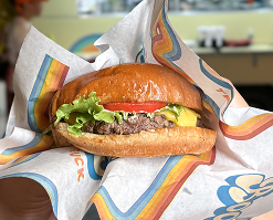 Uma pessoa segura um hambúrguer gourmet em um papel com design do Boogie Truck. O Boogie Truck aparece no fundo da imagem e está à noite.
