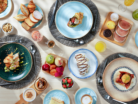 Vista de cima de uma mesa de café da manhã repleta de pratos variados e coloridos, incluindo frutas frescas, pães, bolos, omeletes e sucos.