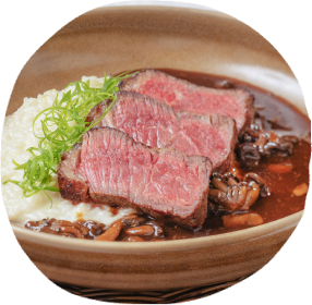 Prato gourmet com cortes de carne, purê e molho, servido em uma tigela de cerâmica. Uma mão segura um copo com um coquetel ao lado, talheres ao lado do prato e um porta tempero ao fundo.