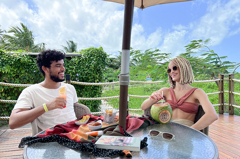 Um casal sorridente desfruta de drinks tropicais, um coco e petiscos em uma mesa de madeira ao ar livre. No fundo há muita vegetação verde e céu azul.