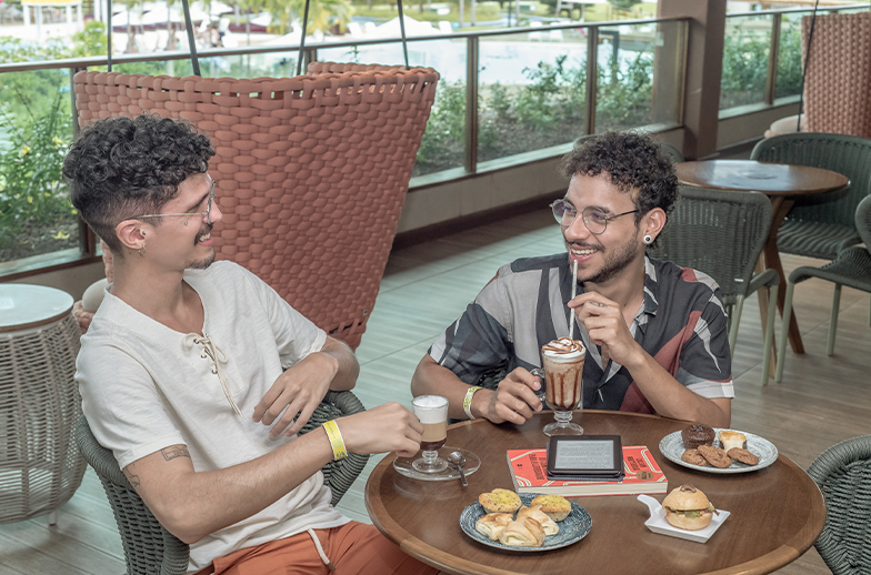 Dois jovens, sorrindo, desfrutam de cafés especiais e petiscos com pães de queijo e doces em uma mesa na área externa. A área da piscina está visível ao fundo.