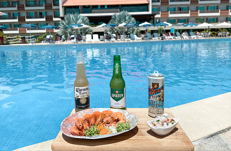 Uma grande variedade de cervejas em garrafas e latas (incluindo Stella Artois, Beck's, Spaten e Corona) dispostas sobre madeira na beira de uma piscina de resort.