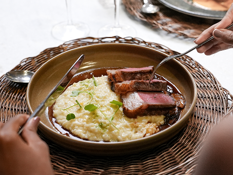 Prato com fatias de carne e um risoto com detalhe de verde e molho da carne, sob um jogo americano de palha. Uma pessoa está com talheres sob o prato.