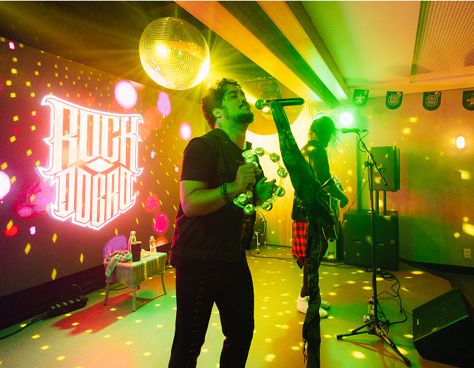 Banda tocando com letreiro ao fundo e uma pequena mesa com bebidas em cima. O cantor está com uma pandeirola e o outro membro toca um violão. Luzes coloridas, e há globo de discoteca pendurado no teto no Pub Overdrive no Japaratinga Lounge Resort.