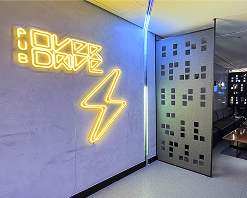 A entrada do Pub Overdrive, com um grande letreiro de neon com a palavra 'OVERDRIVE' e um raio. Uma pessoa está caminhando em direção ao interior do pub, e alguns assentos no caminho até o bar no fundo.