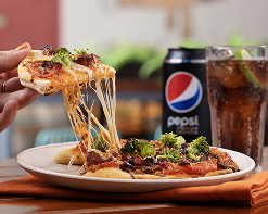 A imagem mostra uma fatia de pizza sendo levantada com uma pá de pizza, com o queijo esticando-se em fios longos e derretidos. Ela tem fatias de proteína e couve-flor. Ela está em prato bege. Há uma lata de pepsi ao lado com copo de gelo com refrigerante e limão dentro.
