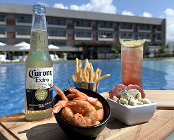 Bandeja com uma garrafa de cerveja, um bowl de camarão e batata frita, e um coquetel, servidos em uma piscina do Bar Verano.
