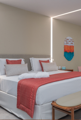 Quarto moderno no Japaratinga Lounge Resort, com uma cama grande, com banco de apoio na frente, iluminação embutida e uma porta de vidro que dá acesso a uma varanda com rede e vista pra as palmeiras.