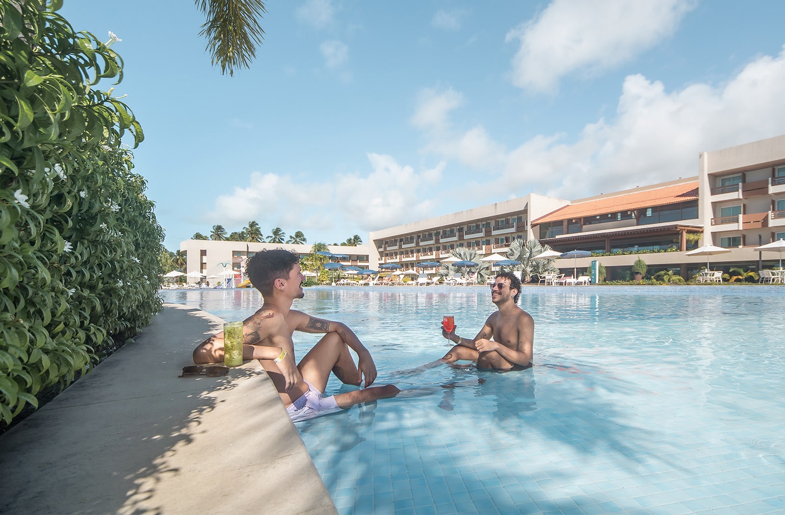  Dois homens sorrindo e relaxando na piscina, um deles sentado na borda e o outro mais dentro da água. Eles seguram drinks, com os prédios do resort e palmeiras ao fundo.