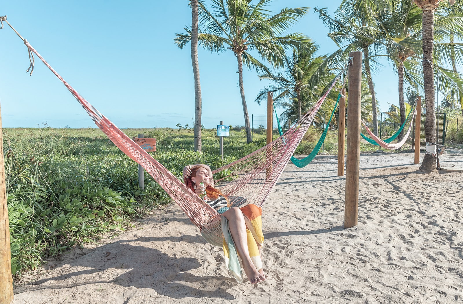 Mulher relaxando em uma rede de balanço, com vários balanços ao redor em uma área de areia, com várias palmeiras e um céu azul claro.