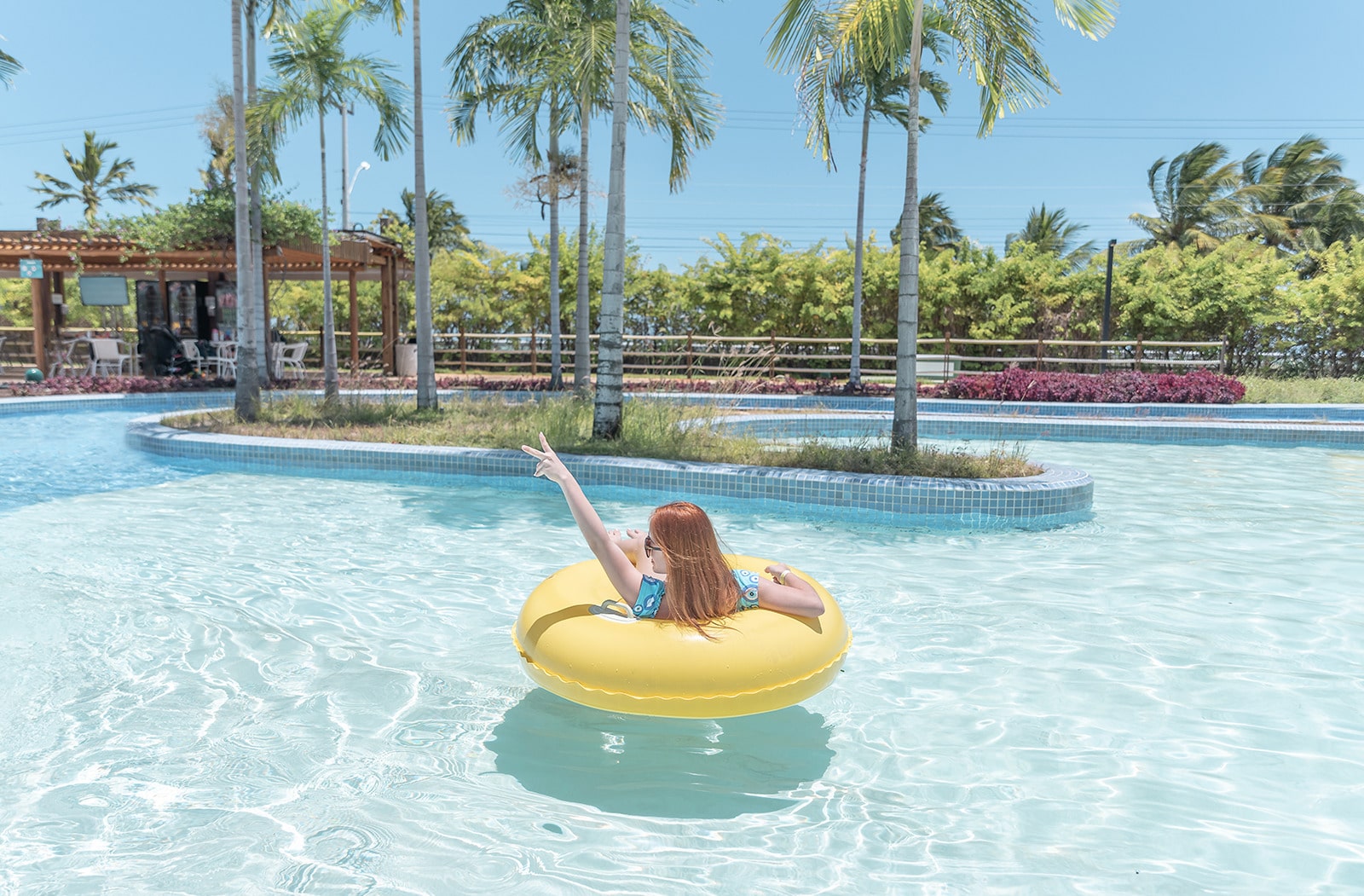 Mulher de costa, deitada em uma bóia amarela, dentro de uma piscina grande. O sol está forte e céu azul sem nenhuma nuvem, com palmeiras e um espaço coberto ao fundo.