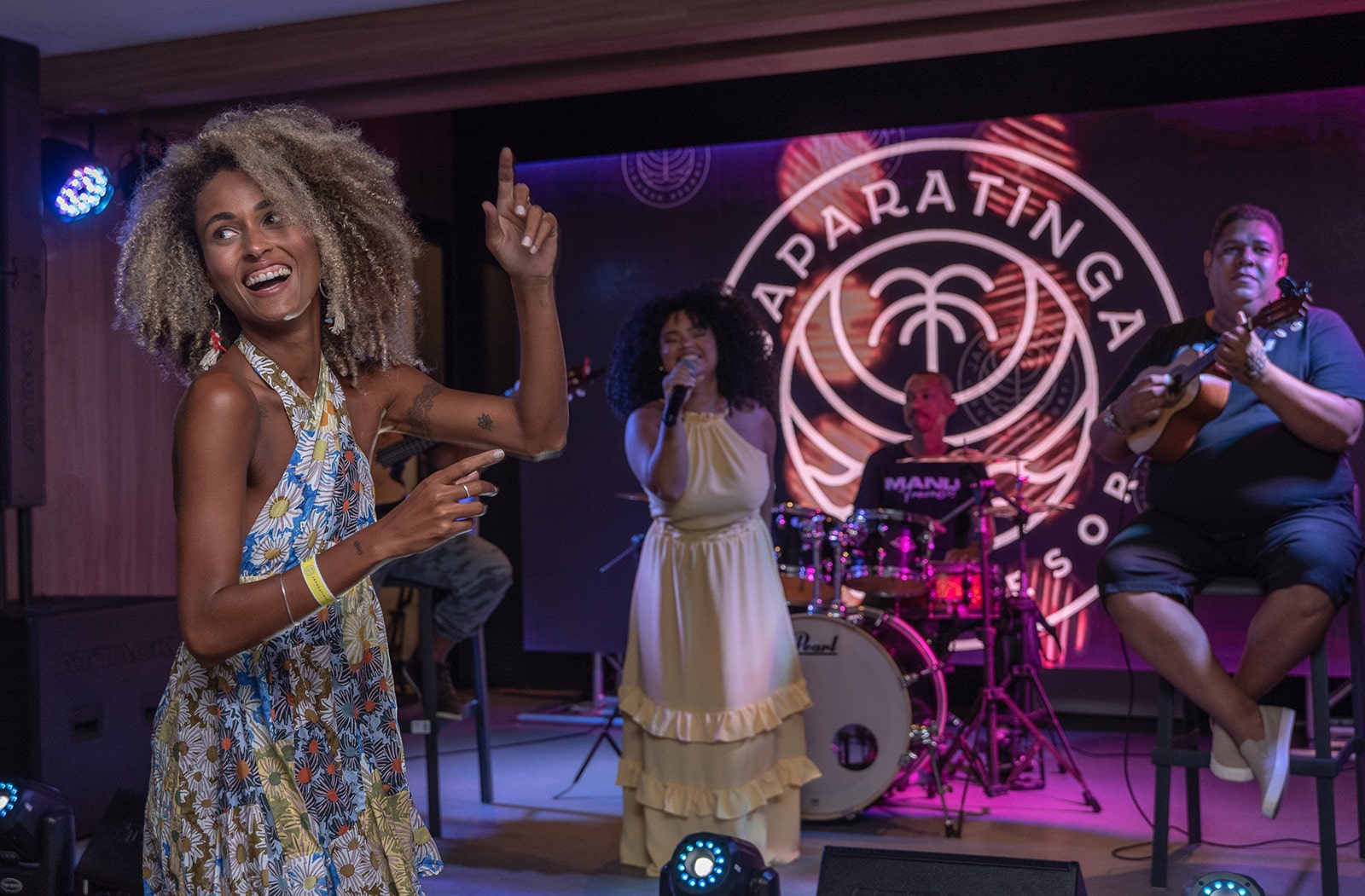 Mulher dançando e sorrindo durante um show de música ao vivo. Uma banda com 4 membros, a cantora é uma mulher com vestido amarelo que se apresenta em um palco com um telão exibindo o logo do resort. O ambiente é iluminado por luzes coloridas.