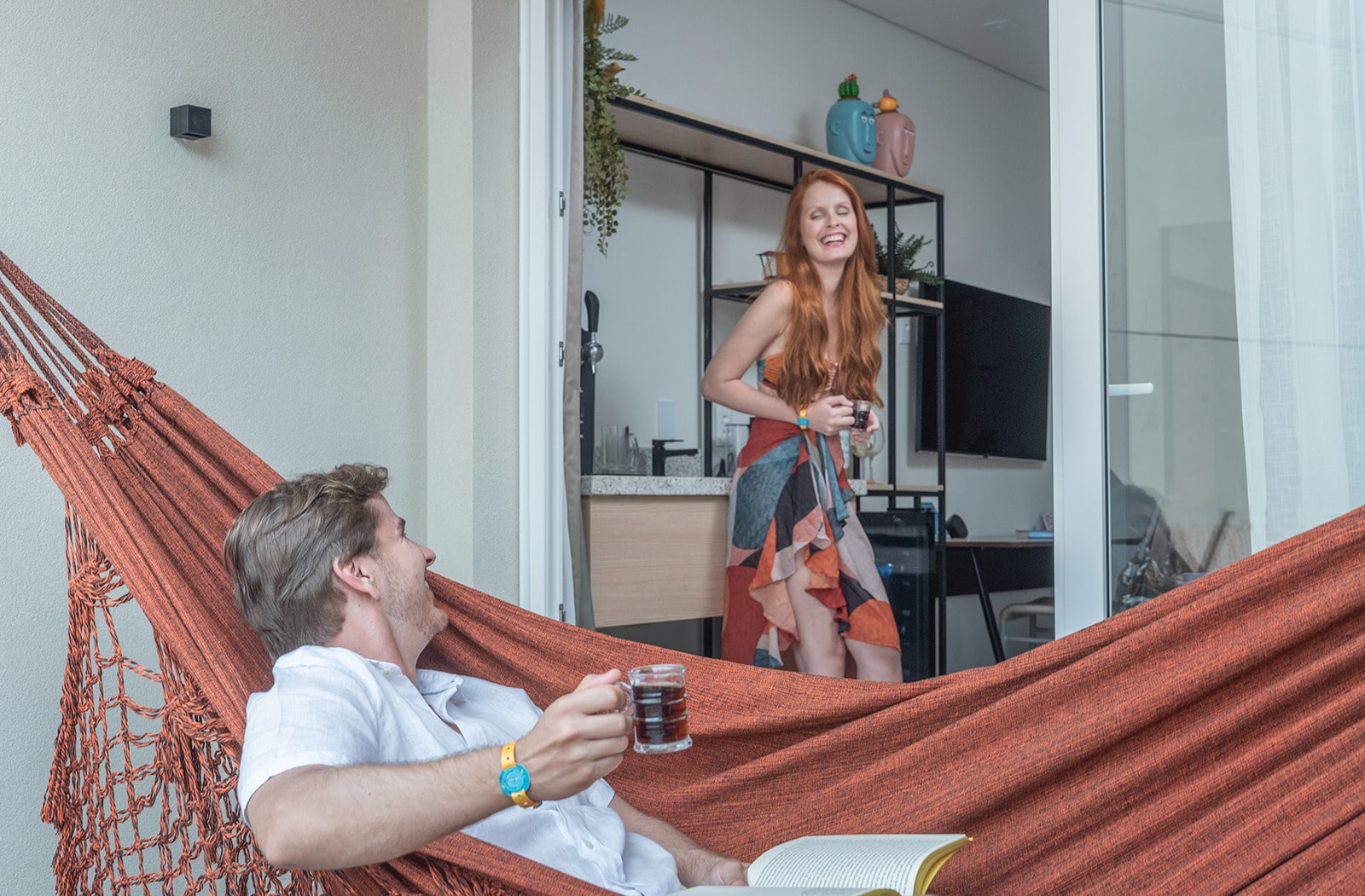 Casal sorrindo e relaxando em uma varanda. O homem está deitado em uma rede com um livro no colo e um drink na mão direita, enquanto a mulher está na porta da varanda do quarto com os móveis e tv no fundo.
