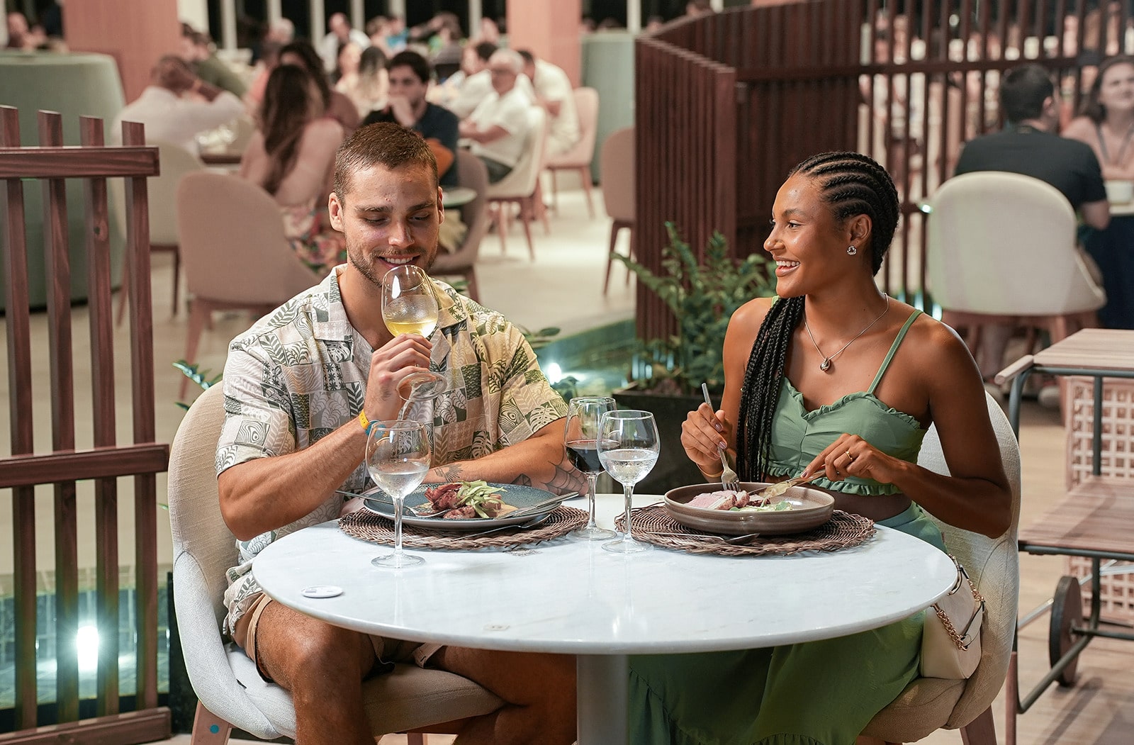 Casal sorrindo e jantando no restaurante Maka com mesas cheias no fundo. O homem segura uma taça de vinho branco e a mulher come sua refeição.