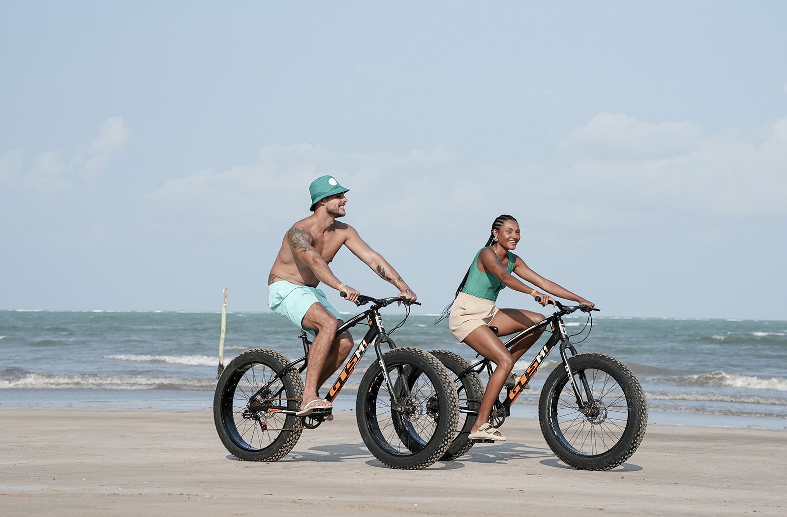Casal pedalando bicicletas com pneus largos na beira da praia. O sol está forte, com o mar em maré baixa e o céu azul ao fundo.