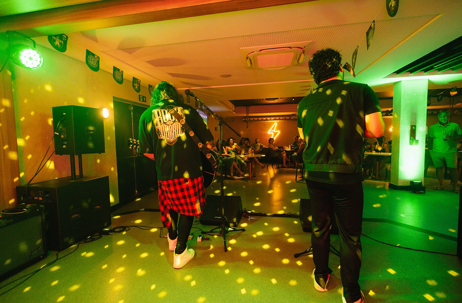 Dois músicos de uma banda tocando em um palco. O público está sentado em mesas no fundo, e o ambiente é iluminado por luzes coloridas. 