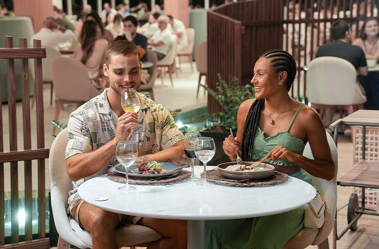  Casal no Restaurante Maka, com pratos de comidas sobre a mesa e duas taças de vinho e duas taças de água, ao fundo o salão do restaurante com hóspedes jantando.