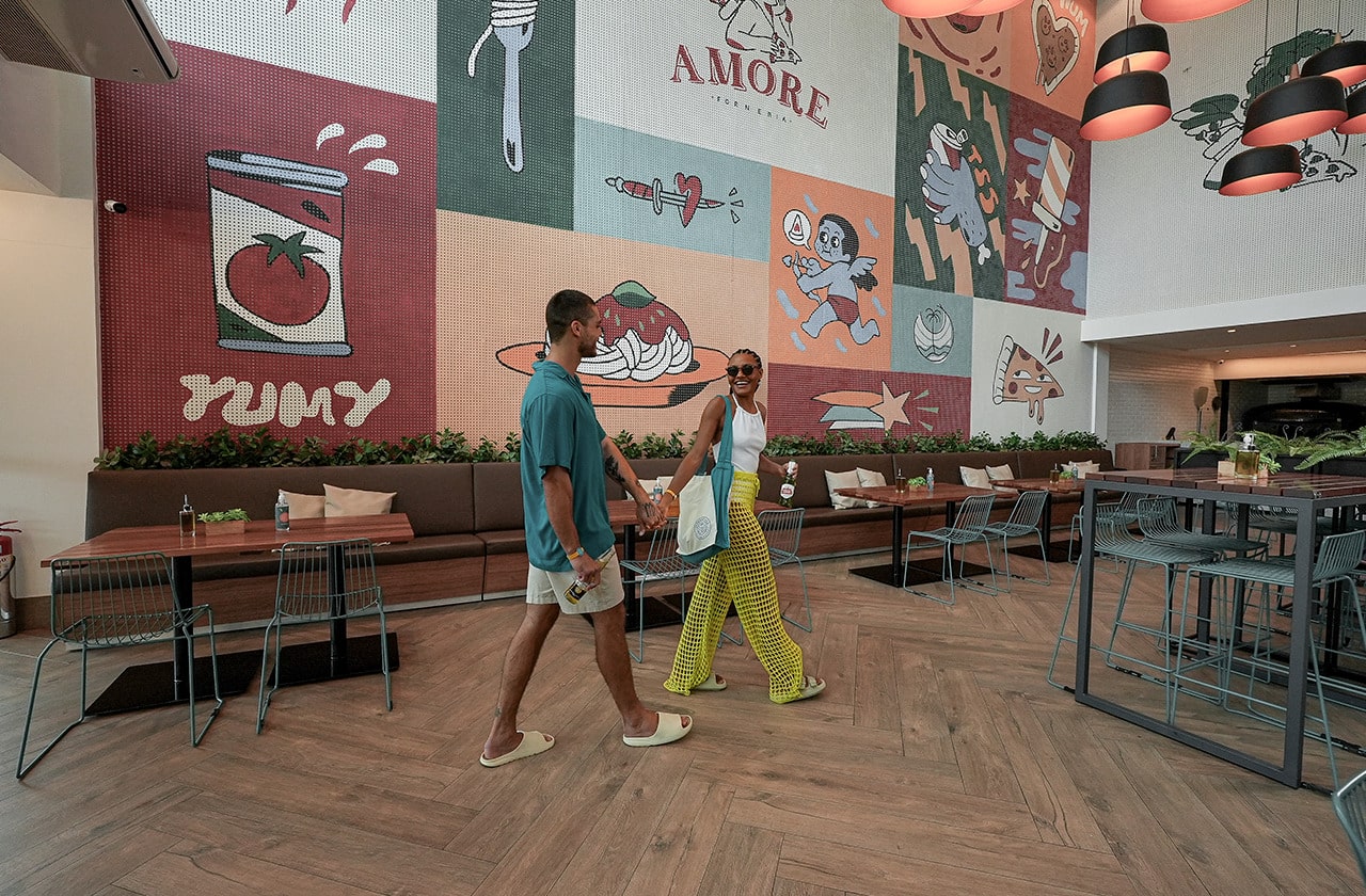 Interior do restaurante Amore Forneria no Japaratinga Lounge Resort, com um casal caminhando no salão. A parede é decorada com um grande mural colorido com ilustrações de pizza e massas e algumas mesas e cadeiras no salão principal.