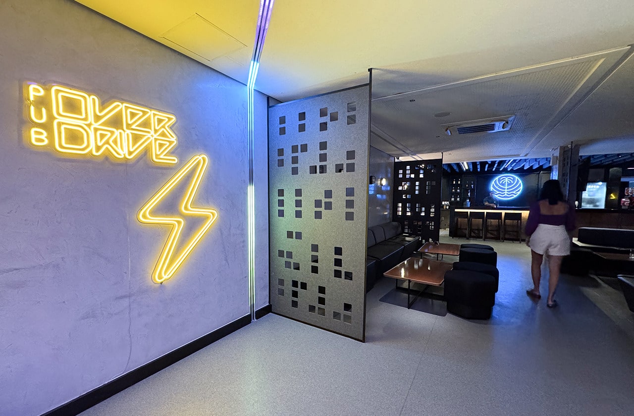 A entrada do Pub Overdrive no Japaratinga Lounge Resort, com um grande letreiro de neon com a palavra 'OVERDRIVE' e um raio. Uma pessoa está caminhando em direção ao interior do pub e alguns bancos no caminhos