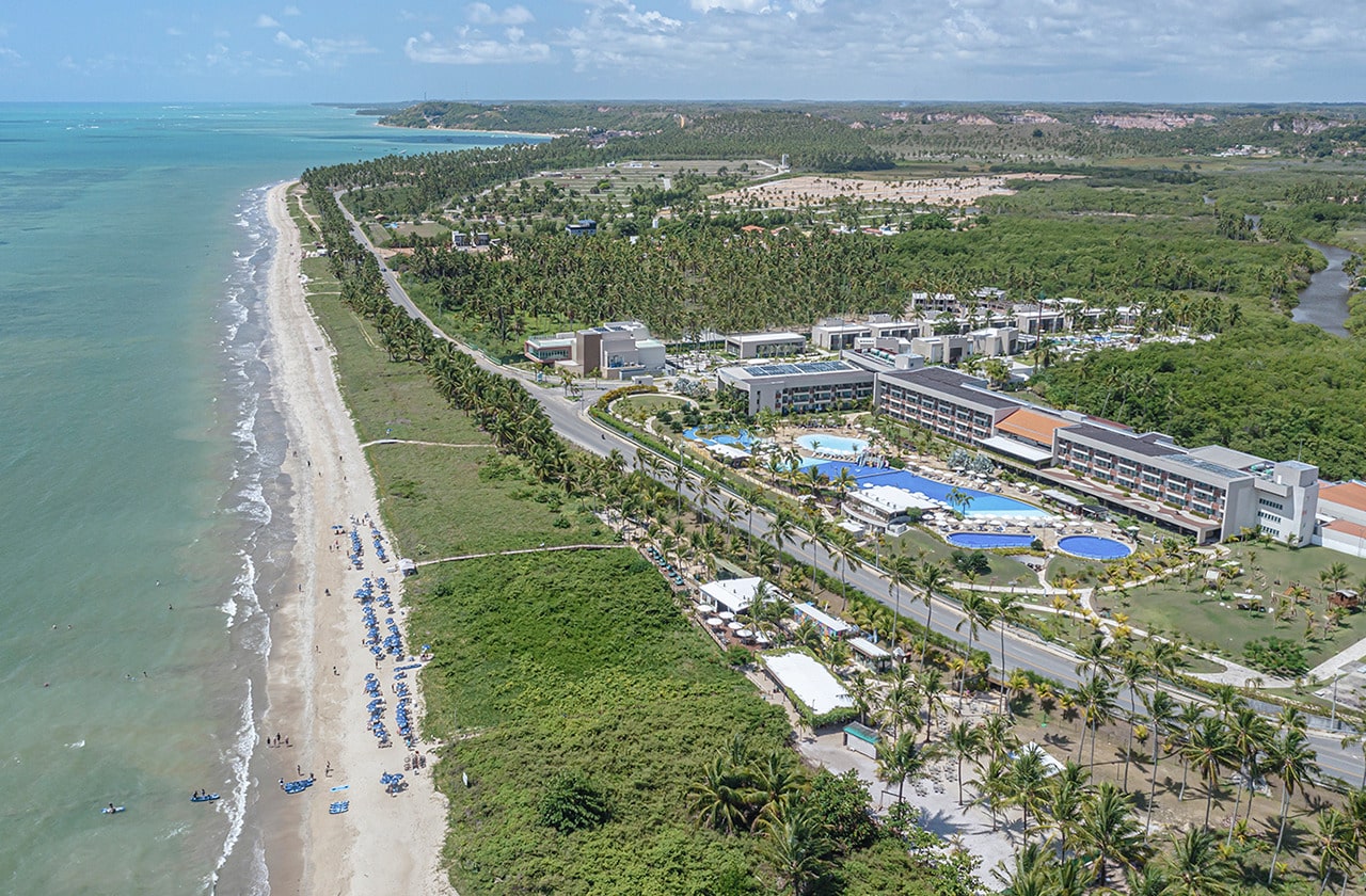 Vista aérea do Japaratinga Lounge Resort, mostrando o complexo ao lado da praia com água azul, com coqueiros, faixa de areia, piscinas e a natureza paradisíaca verde do local.