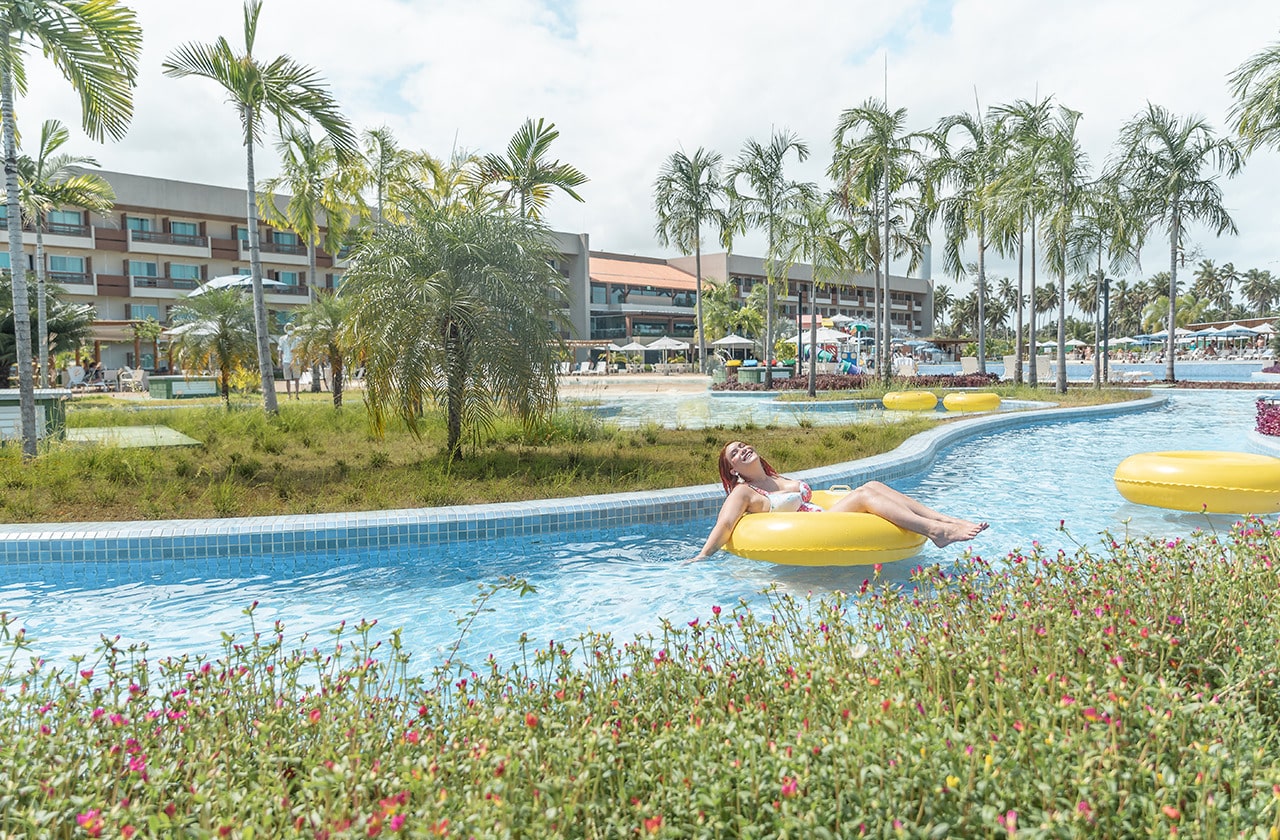 Mulher relaxando em uma bóia amarela na piscina de correnteza do Japaratinga Lounge Resort, com os apartamentos ao fundo e bastantes verdes das plantas.