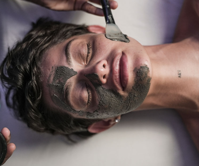 Uma mulher está recebendo um ritual facial, com uma máscara de argila no rosto sendo aplicada com um pincel por uma pessoa. Os olhos estão fechados e o ambiente é de relaxamento, mostrando o tratamento de spa.